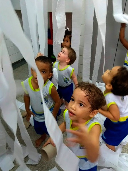 Em sala de aula, cinco crianças uniformizadas, com camisas brancas e shorts azuis, olham admiradas para rolos de papel higiênico suspensos e presos a cordas horizontalizadas. Fim da descrição