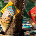 Livros pendurados em árvore com fitas coloridas. Em volta da árvore, dezenas de livros sobre mesas. Fim da descrição.