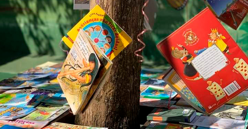 Livros pendurados em árvore com fitas coloridas. Em volta da árvore, dezenas de livros sobre mesas. Fim da descrição.