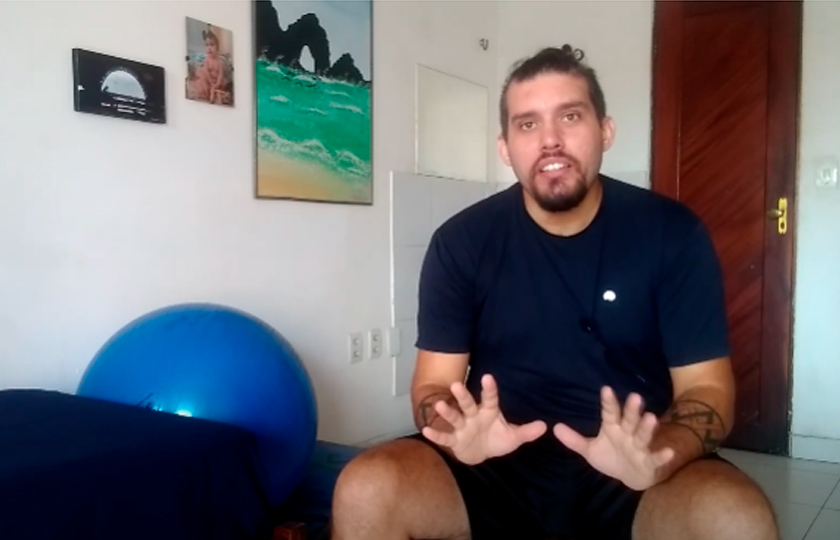 Captura de tela de educador André Cyrino falando com seus estudantes em vídeo. Ele usa roupa preta e está sentado em banco em frente à câmera. Ao lado, aparece um sofá e uma bola azul de ginástica. Fim da descrição.
