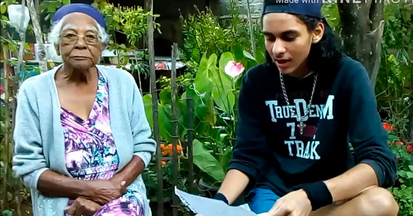 Jovem e sua avó sentados lado a lado. O estudante lê algo de papel em sua mão. A idosa olha para a câmera. Fim da descrição.