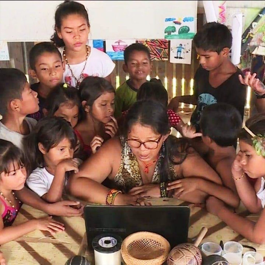 Sentada em cadeira e em frente a notebook, Claudia Baré é rodeada por pequenos estudantes indígenas. Ela usa óculos e pulseiras nos braços. Fim da descrição.
