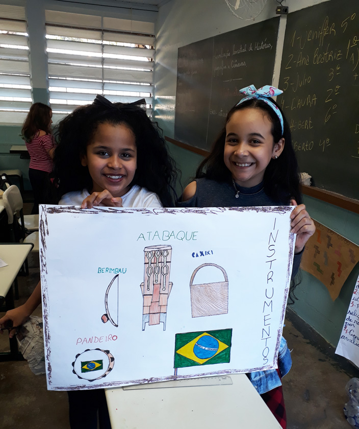 Em sala de aula, duas alunas sorridentes seguram cartaz sobre instrumentos musicais de origem africana. É possível observar com imagens e palavras o atabaque, o pandeiro, caxixi e o berimbau, além de uma bandeira do Brasil desenhada. Fim da descrição.