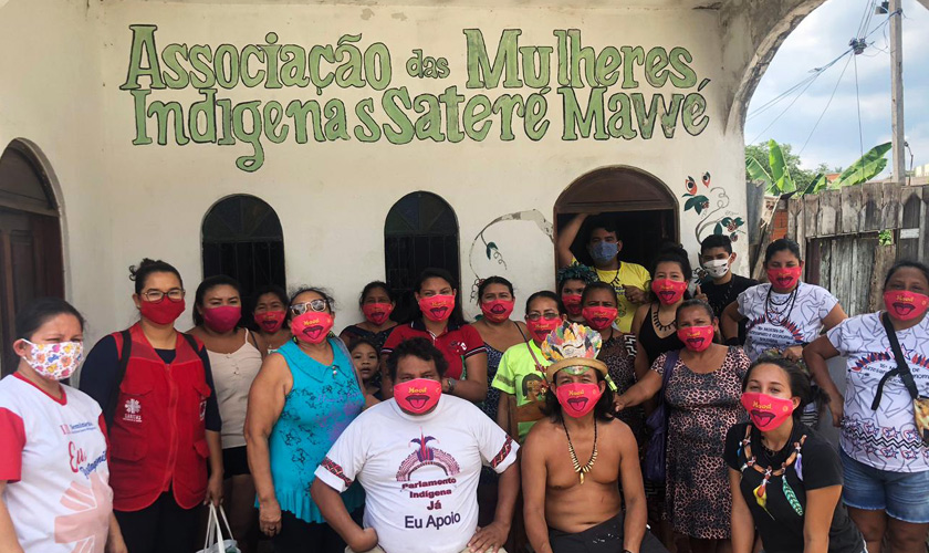 Cerca de vinte indígenas posam para foto utilizando máscaras vermelhas. Alguns deles usam adornos tradicionais. Ao fundo, parede bege escrito em verde "Associação das Mulheres Indígenas Sateré Mawé". Fim da descrição.