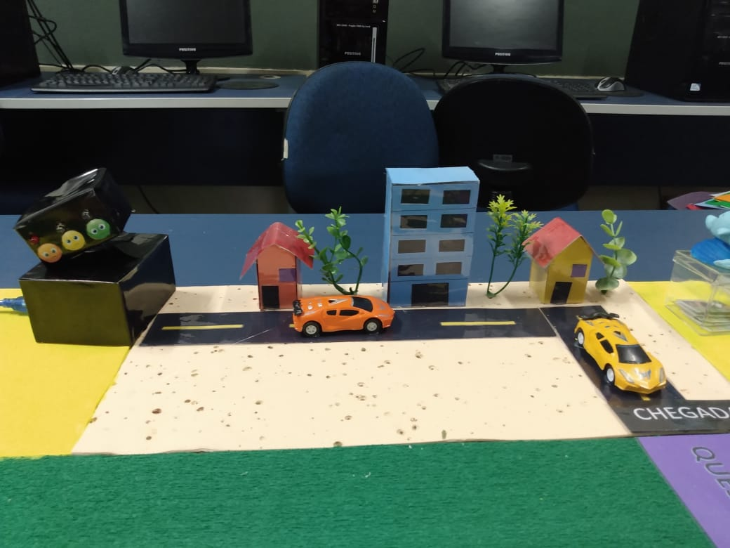 Decoração do tabuleiro com carrinhos de brinquedo, e prédios e árvores de papel. Fim da descrição.