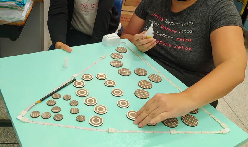 Duas educadoras realizam a montagem do jogo "A teia". Uma dela segura uma placa de madeira em verde claro, enquanto a outra posiciona círculos de diferentes tamanhos dentro de um retângulo posicionado no painel de madeira. Fim da descrição.