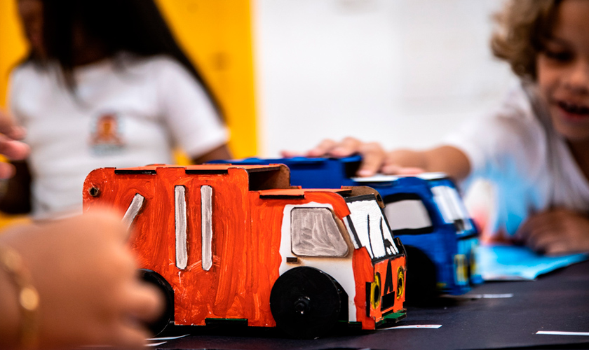 Em primeiro plano, caminhão laranja está parado no Caminho sustentável. Ao fundo, estudante está com a mão sobre um caminhão azul. Fim da descrição.