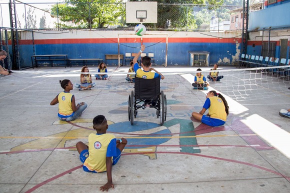 Crianças jogam vôlei sentados na quadra. Ao centro, um menino em cadeira de rodas se prepara para rebater a bola que chega até ele. Fim da descrição.