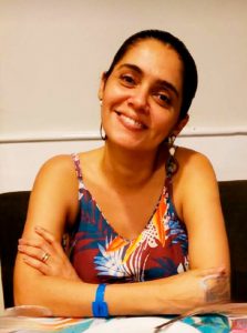 Educadora Helayne Carvalho, mulher com cabelo escuro, sorri para a foto. Ela está com o cabelo preso e usa brincos e blusa floral.