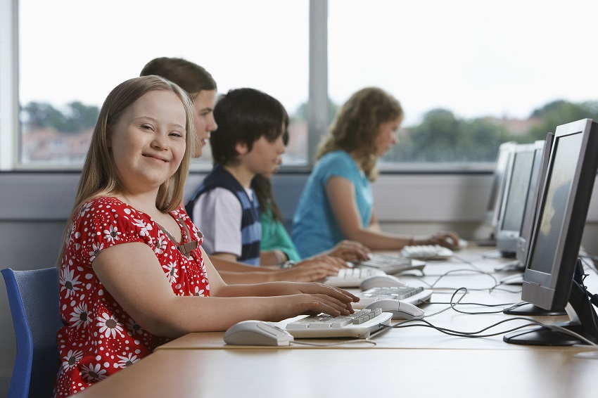 Sentados à mesa um ao lado do outro, cinco adolescentes utilizam computadores em sala de aula. Fim da descrição. 