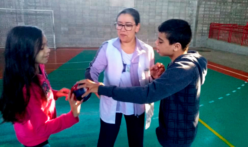Em quadra esportiva, guia intérprete Valdineia Nascimento acompanha estudante surdocego em jogo com outra estudante.