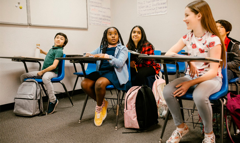 Em sala de aula, cinco estudantes de diferentes etnias participam de aula. Uma das alunas fala e os outros sorriem.