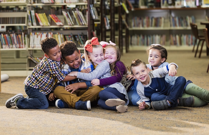 Sentados no chão da biblioteca, seis crianças sorriem e se abraçam. Fim da descrição. 