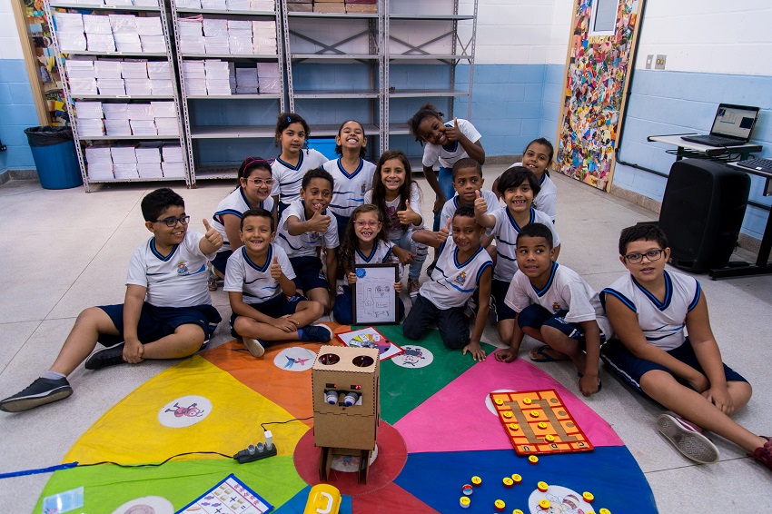 Em sala de aula, estudantes estão sentados no chão em volta de tapete colorido, posando para foto. O material pedagógico acessível Arizinho está no centro. 