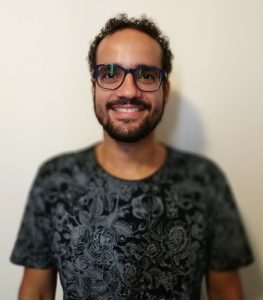 Orientador pedagógico, Tiago Ribeiro sorri para a câmera. Ele é um homem adulto, com cabelo curto enrolado, barba e óculos de grau. Fim da descrição.
