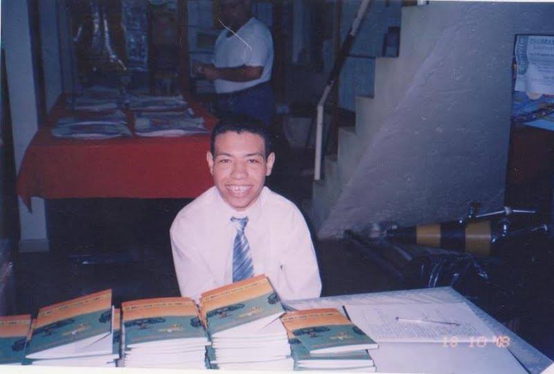 Foto de 2008. José Roberto está atrás de mesa com vários exemplares de seus livros empilhados. Ele está de camisa social e gravata e sorri.