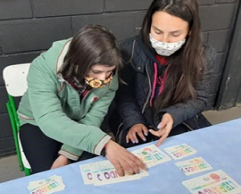 Estudante está sentada participando do jogo Librando, com o auxílio de educadora. Fim da descrição.