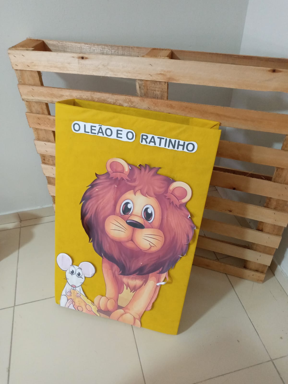 Livro amarelo, com o título o leão e o ratinho e a figura dos dois animais, está em pé escorado em um pallet de madeira. Fim da descrição. 