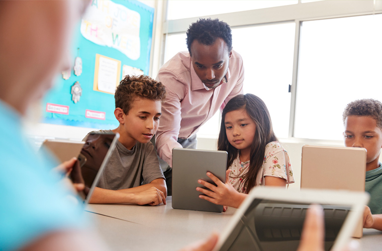 Três estudantes, dois meninos e uma menina, com tablets enquanto educador os orienta em sala de aula. Fim da descrição.