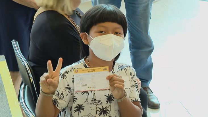 Criança indígena utilizando máscara posa para foto com comprovante de vacinação na mão esquerda, enquanto faz sinal de paz com a mão direita. Fim da descrição.