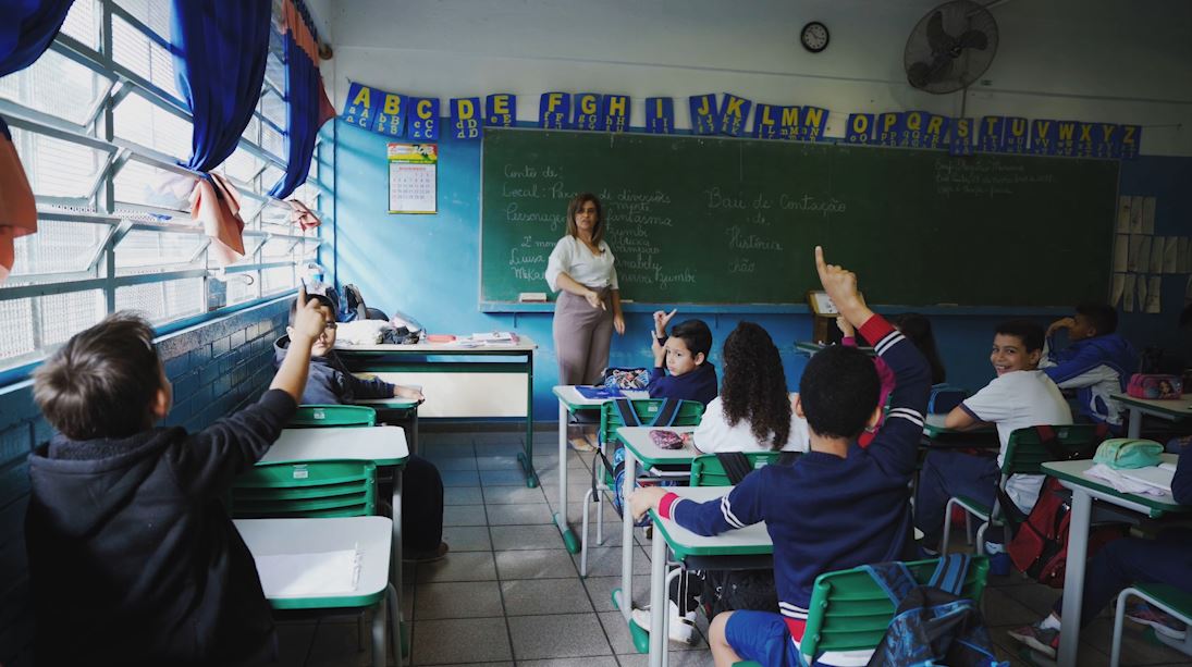 Em sala de aula, estudantes estão sentados nas carteiras. Três deles levantam a mão enquanto professora os observa em pé em frente à lousa. Fim da descrição.