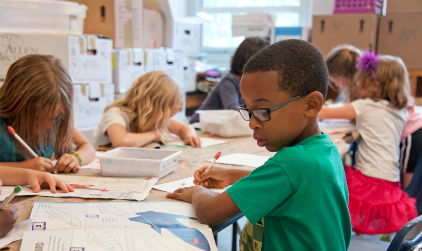 Em sala de aula, crianças estão sentadas em mesas conjuntas e escrevem em livros. Mais próximo da câmera, menino de óculos observa figura em livro, enquanto escreve em outra folha. Fim da descrição.