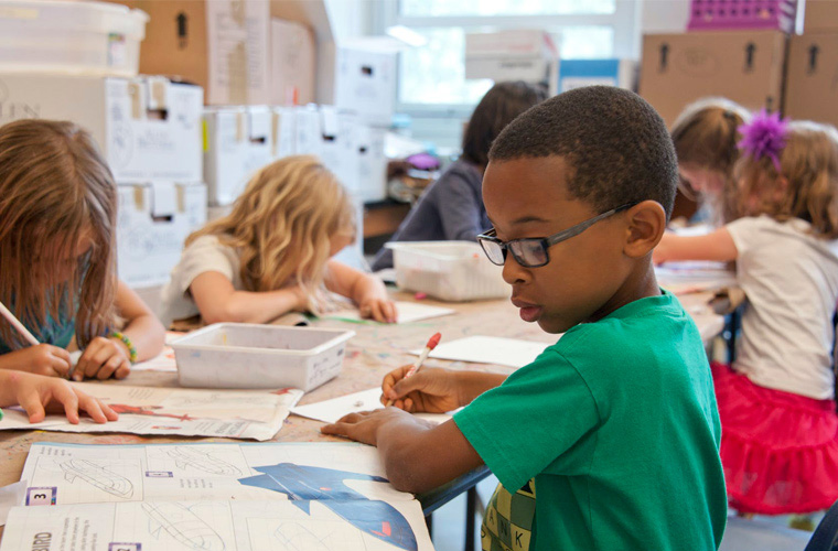 Em sala de aula, crianças estão sentadas em mesas conjuntas e escrevem em livros. Mais próximo da câmera, menino de óculos observa figura em livro, enquanto escreve em outra folha. Fim da descrição.