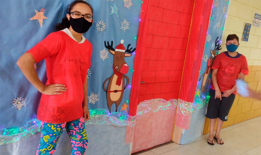 Em corredor de escola, dois estudantes, um menino e uma menina, estão ao lado de pinturas de natal na parede. Ambos usam máscara. Fim da descrição.