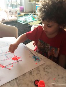 Em casa, menino utiliza pincel e tinta vermelha para fazer atividade escolar. Fim da descrição.