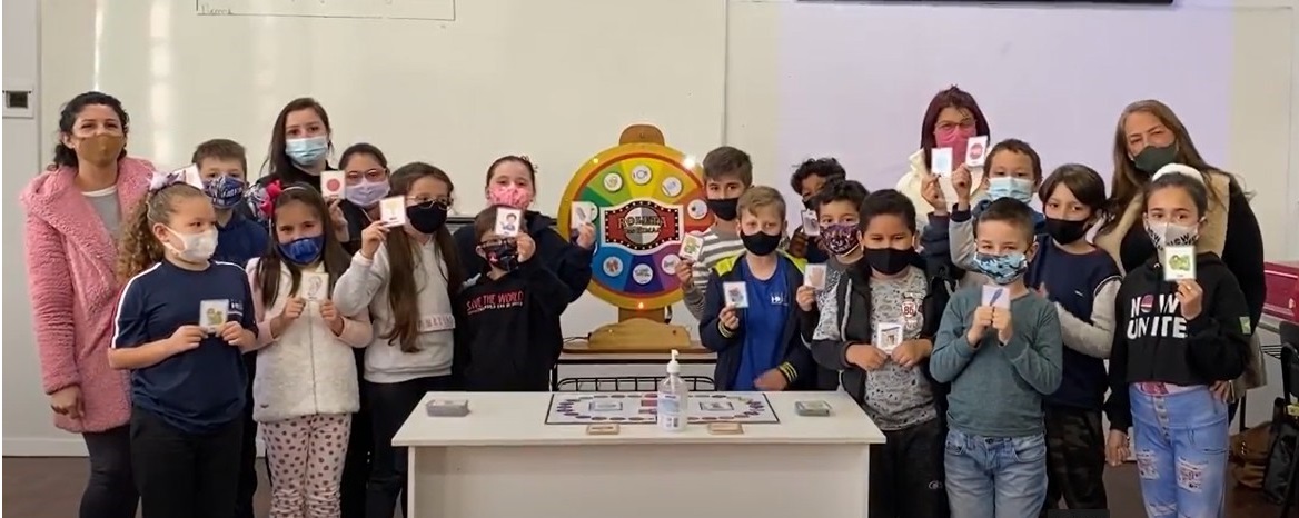Estudantes e educadoras posam para foto reunidos em volta do material pedagógico acessível com cartas do jogo nas mãos. Todos usam máscara. Fim da descrição.