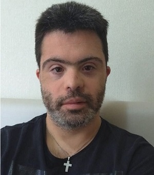 João Vitor posa para foto usando uma camiseta preta e uma corrente no pescoço, com pingente prata e formato de cruz. Ele é um homem branco na faixa etária de 30 anos, com barba, bigode e cabelos grisalhos. Fim da descrição.