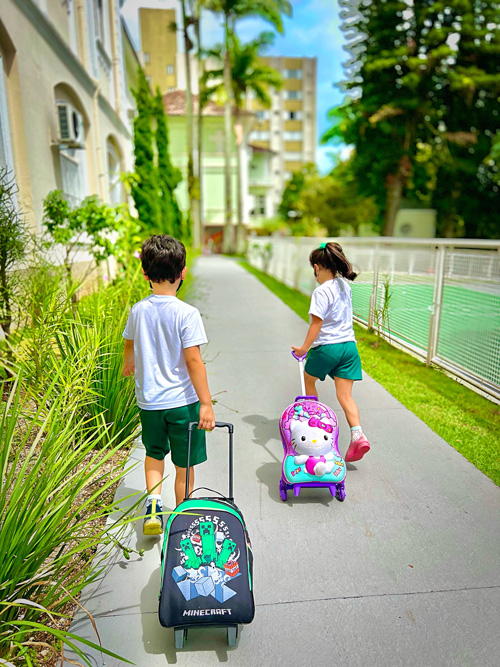 Menino e menina, uniformizados e carregando mochila de rodinhas, aparecem de costas, andando em passarela com plantas e grama ao redor. Fim da descrição.