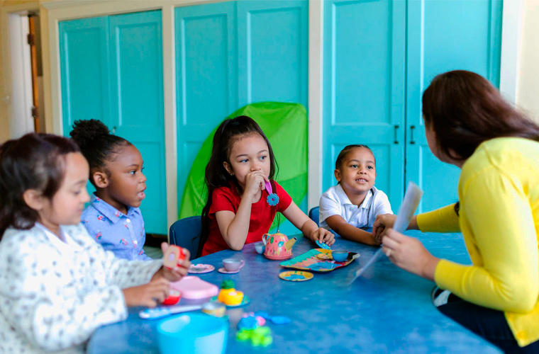 Quatro meninas estão sentadas ao redor de mesa com educadora. Elas mexem com peças coloridas, enquanto educadora mostra um papel. Fim da descrição.