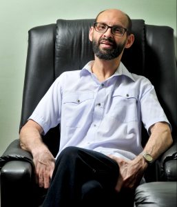 Emílio Figueira, homem com barba, camisa social e óculos de grau, está sentado em poltrona preta, com as pernas cruzadas. Fim da descrição. 
