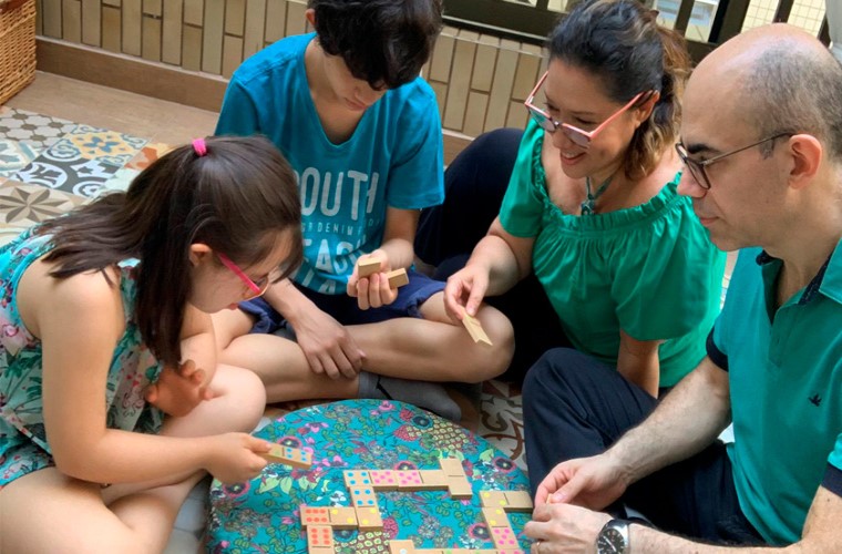 Em espaço externo, mãe e pai jogam dominó com os filhos, um menino e uma menina. Fim da descrição.