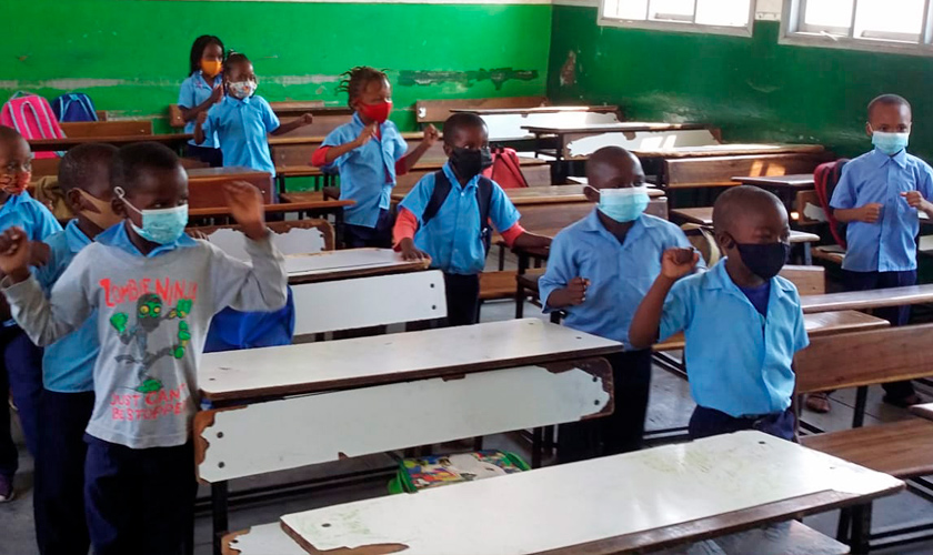 Em sala de aula, crianças uniformizadas estão em pé ao lado de suas carteiras. Todas usam máscara cobrindo o nariz e a boca. Fim da descrição.