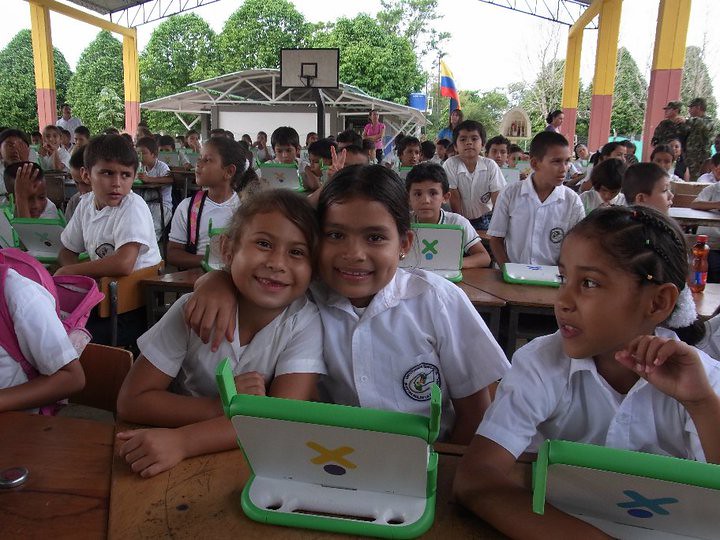 Duas meninas sorriem para foto, sentadas em carteiras em espaço externo da escola com demais estudantes. Fim da descrição.   