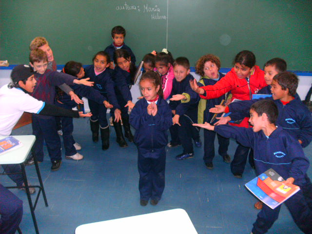 Em sala de aula, uma menina está em pé e ao seu redor outros estudantes esticam a mão em sua direção. Fim da descrição.