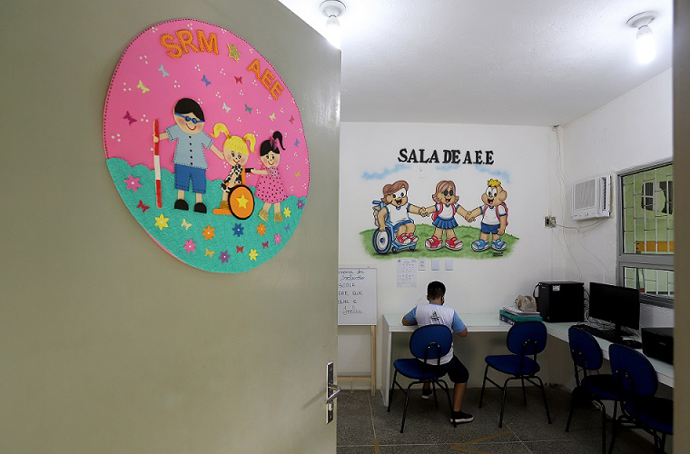 Imagem mostra a sala de recursos multifuncionais vista da porta, onde estudante está sentado durante aula. Fim da descrição.