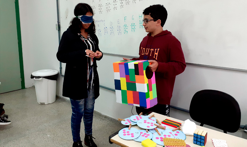Em sala de aula, uma estudante do curso “Braille básico” está vendada enquanto um colega segura caixa colorida com entradas circulares nas laterais. Fim da descrição.