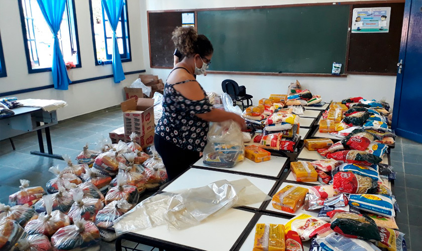 Carteiras escolares estão juntas no centro de sala de aula. Em cima das mesas, alimentos embalados. Educadora os recolhe para montar kits dentro de sacolas transparentes. Atrás dela, no chão, dezenas de kits de alimentos prontos. Fim da descrição.