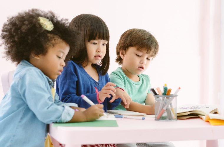 Uma menina asiática, uma negra e um menino branco estão pintando cadernos que estão na mesa. Fim da descrição.