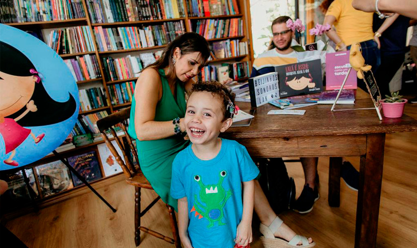 Em biblioteca, menino utilizando implante coclear sorri para a câmera. Ao fundo, estantes de madeira com diversos livros e mesa com uma escritora autografando exemplares de livros infantis. Fim da descrição.