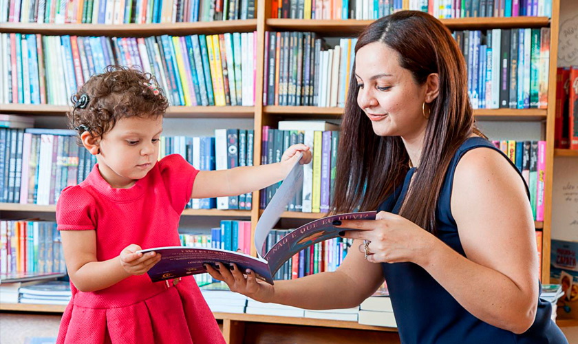 Em biblioteca, menina utilizando implante coclear abre livro com apoio de mulher adulta. Ao fundo, estantes de madeira com diversos livros. Fim da descrição.