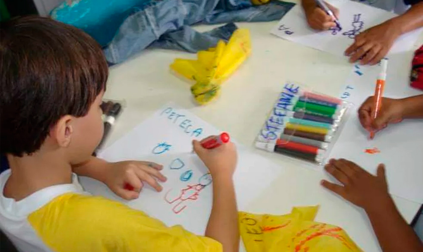 Em sala de aula, três estudantes estão ao redor de mesa quadrada, usando canetinhas coloridas para desenhar em papel branco. Fim da descrição.