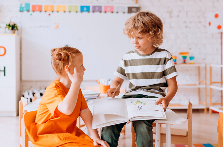 Em sala de aula, uma menina e um menino olham um para o outro enquanto menina faz sinal com um dos dedos em sua cabeça e o menino segura um livro aberto. Fim da descrição.