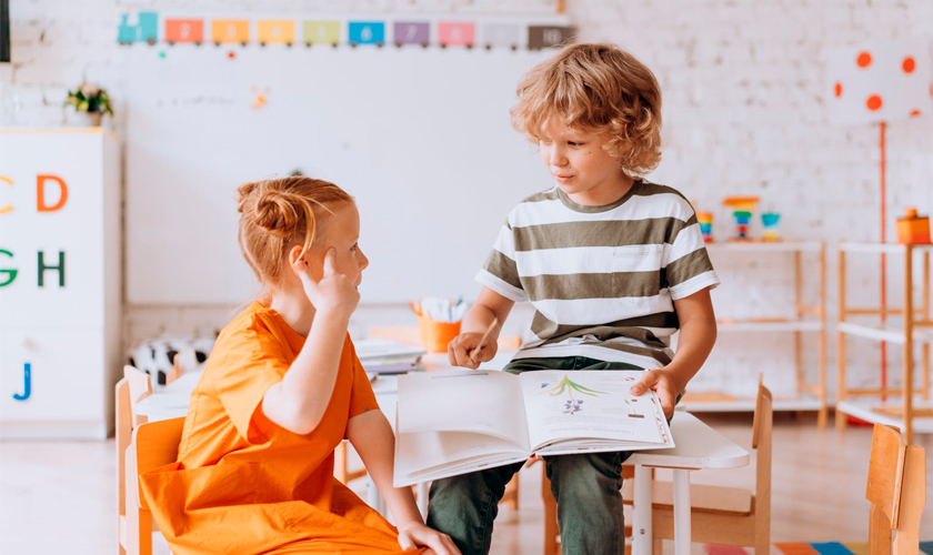 Em sala de aula, uma menina e um menino olham um para o outro enquanto menina faz sinal com um dos dedos em sua cabeça e o menino segura um livro aberto. Fim da descrição.