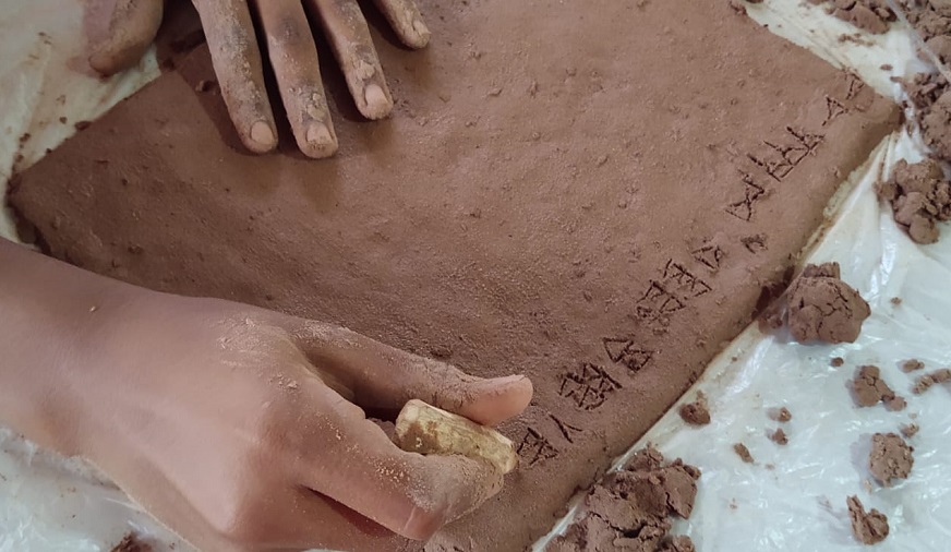 Imagem mostra mãos de estudantes realizando a escrita cuneiforme em argila. Fim da descrição.