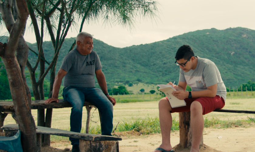 Em campo aberto, estudante e seu pai estão sentados em bancos de madeira, o menino segura um caderno e uma caneta. Fim da descrição.