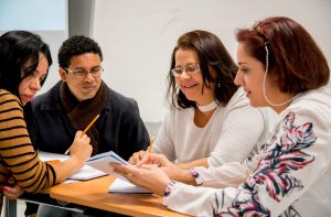 Três professoras e um professor estão sentados à mesa e observam anotações em um caderno. Fim da descrição.